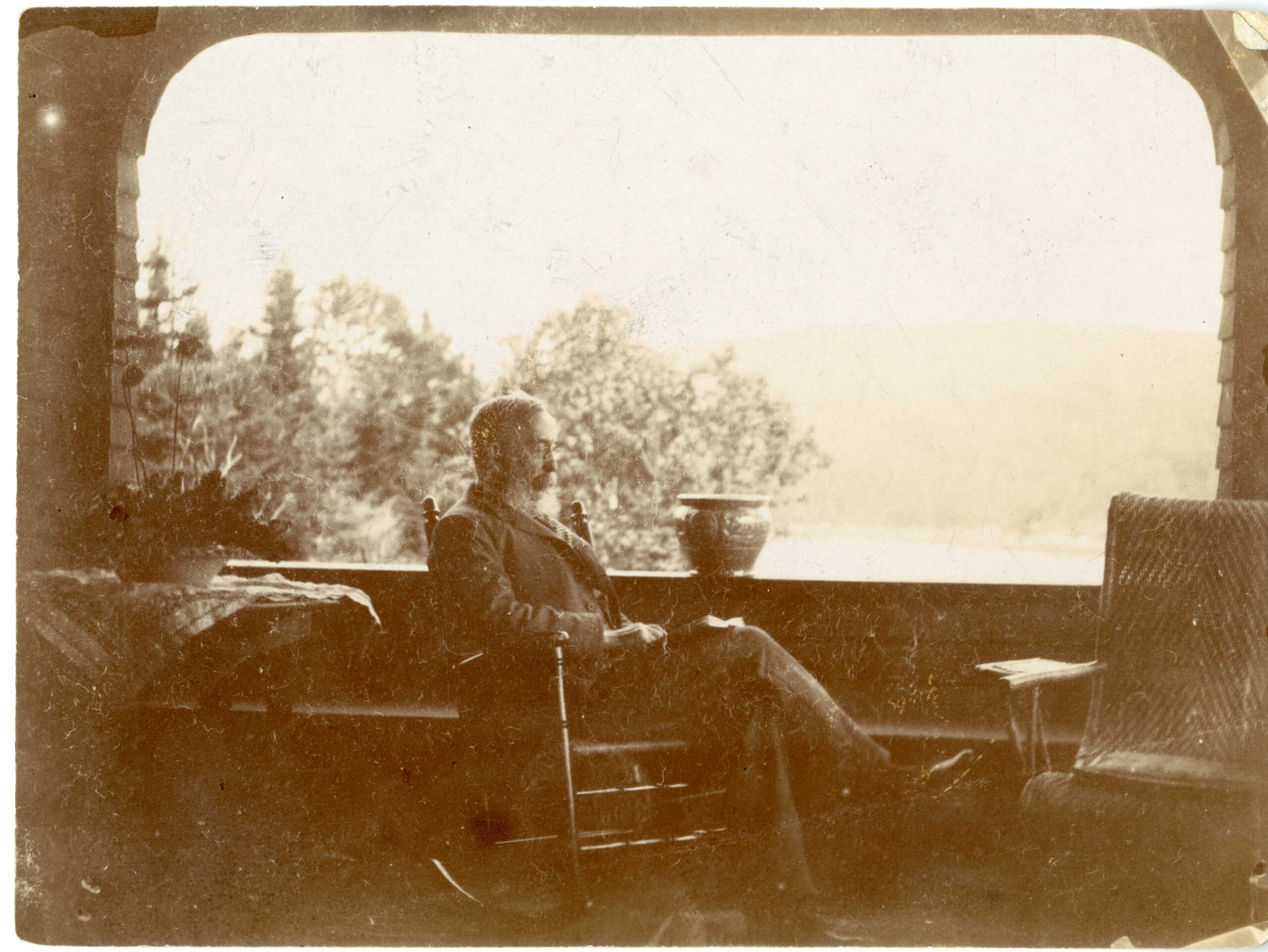 Candid photograph of Gilman at approximately 75 years old reading in a rocking chair on his porch in Maine