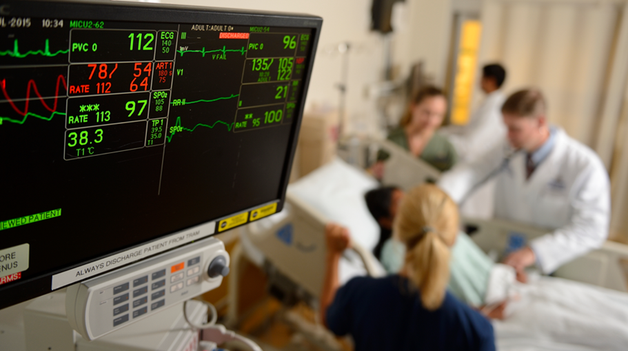 A computer screen shows a patient's vital signs. Blurred in the background, you can see a care team at a patient's bedside.