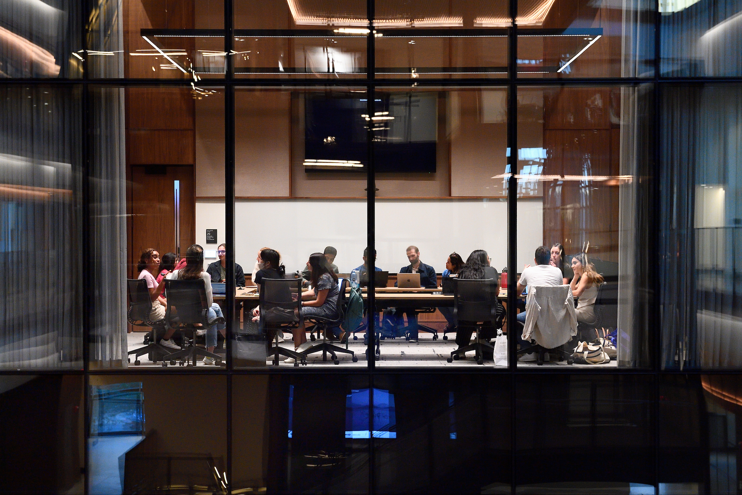 Looking through the window of a classroom, you can see students sitting around a conference table.
