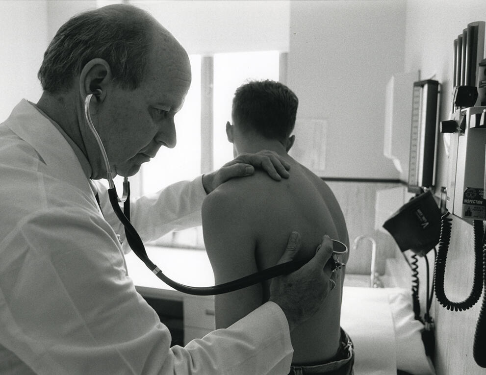 In this black and white photo, a doctor presses a stethoscope to back of a shirtless patient seated on an examination table.