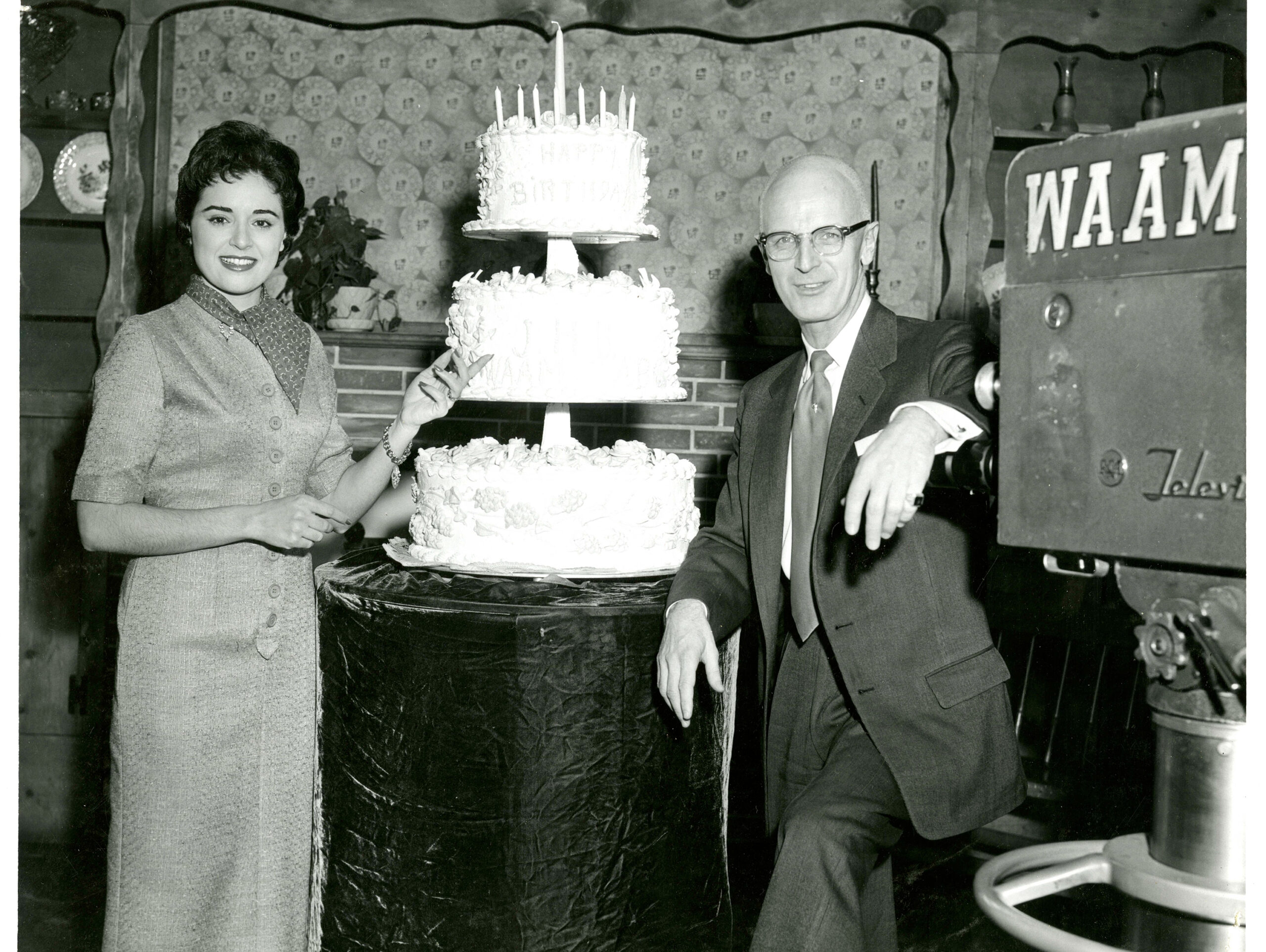 In this black and white photo, a man and woman stand next to a large, three-tiered white cake