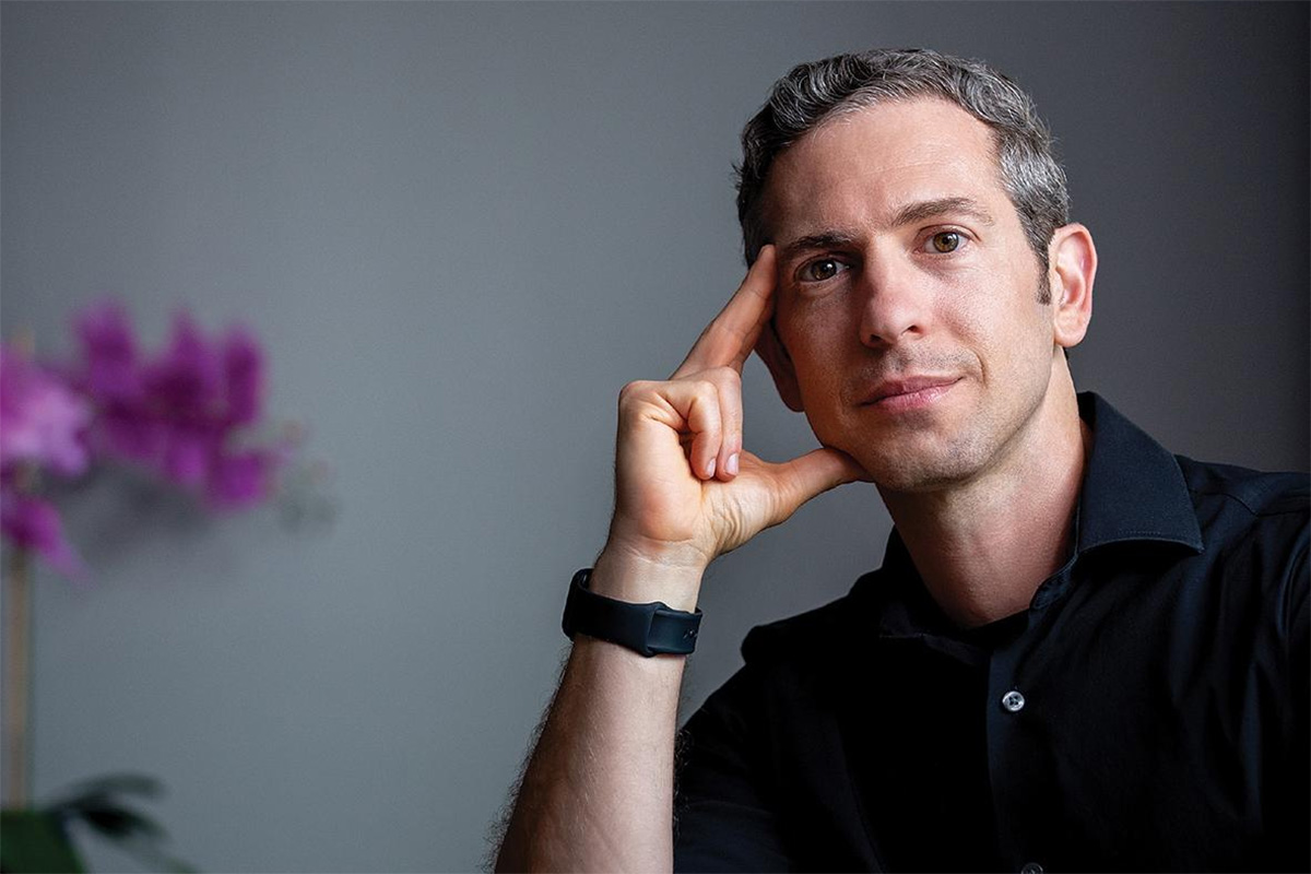 A man in a block shot poses for a portrait; he leans his head on his right thumb and forefinger
