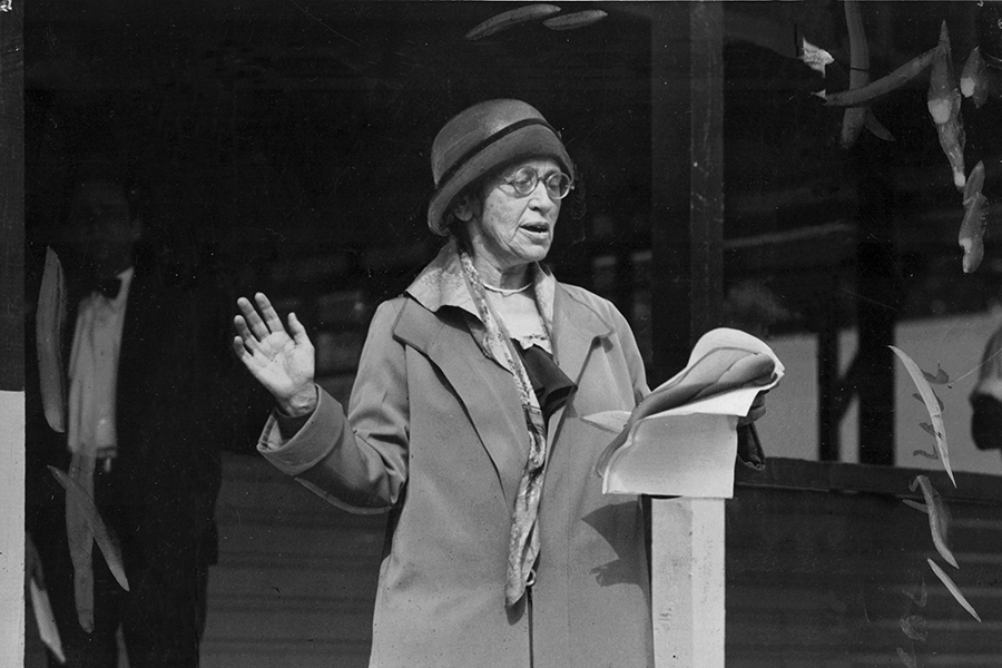 In this black and white photo, a woman in a hat and coat stands and reads aloud from a manuscript held in her left hand