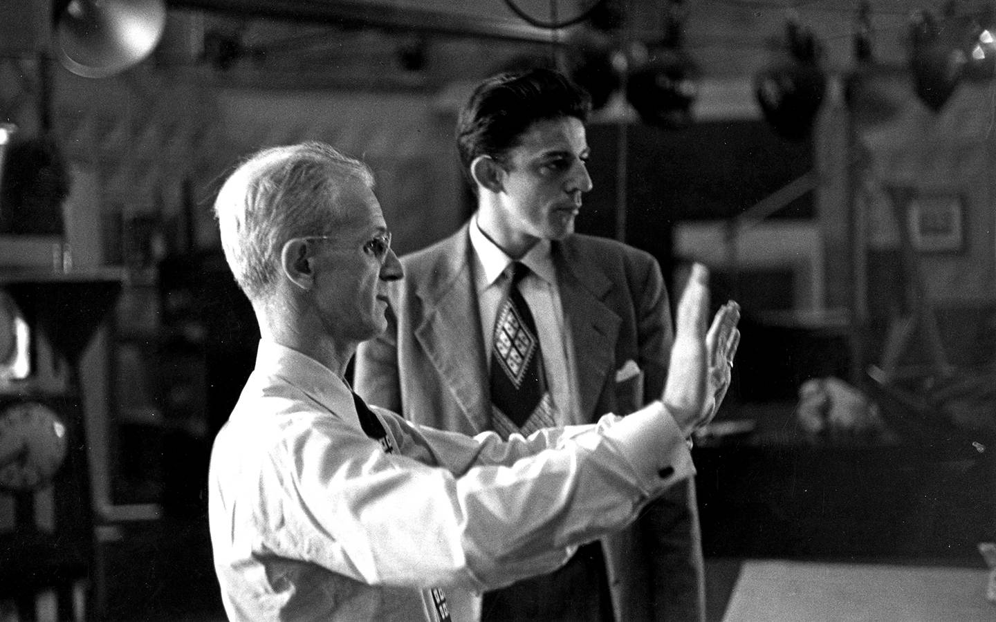 In this black and white photo, a man spreads his hands out in front of him, giving stage directions to another man standing next to him